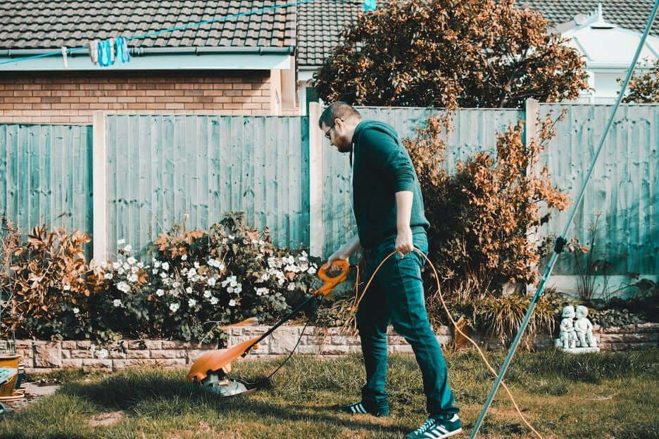 Man mowing the lawn with an electric trimmer in a garden, surrounded by flowers and a wooden fence.