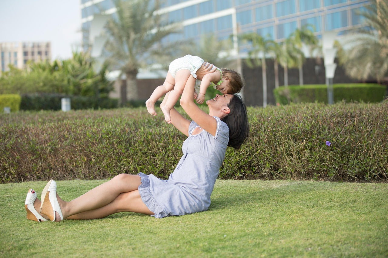 Mother joyfully lifting her baby in a park, surrounded by greenery and modern buildings.
