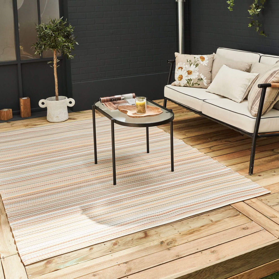 Beige outdoor rug with colorful stripes on a wooden patio, featuring modern decor with a sofa and a round table.