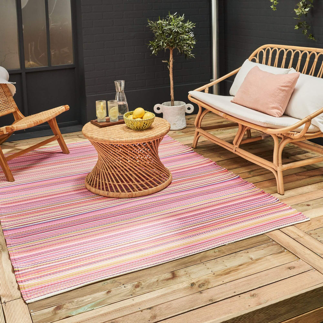 Pink Rainbow Outdoor Rug with colorful stripes on a wooden deck, enhancing a cozy patio setting with wicker furniture and potted plant.