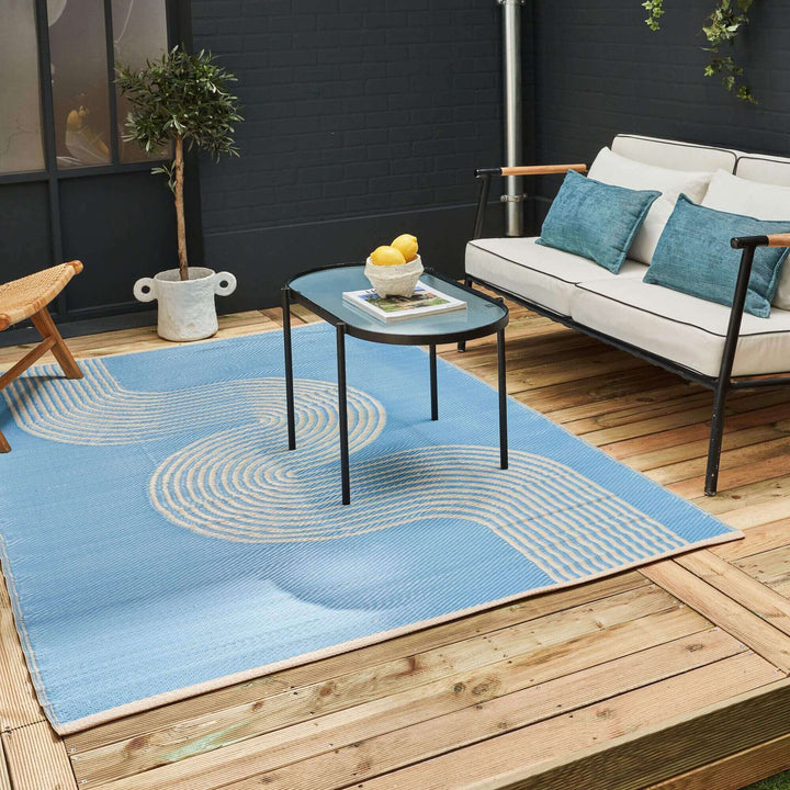 Blue and beige geometric outdoor rug on a wooden patio with modern furniture and potted plant.