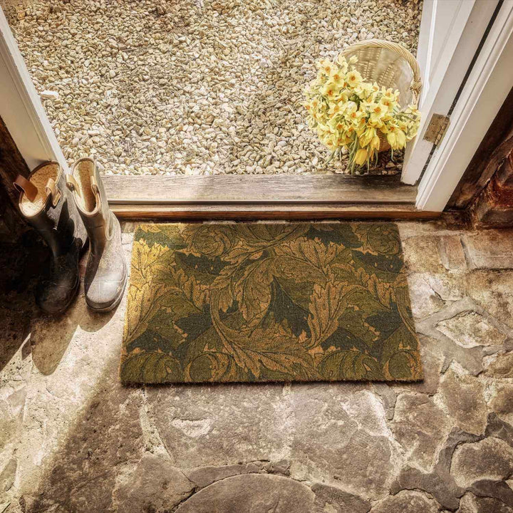 Doormat with leaf pattern on stone floor, shoes, and basket of flowers.