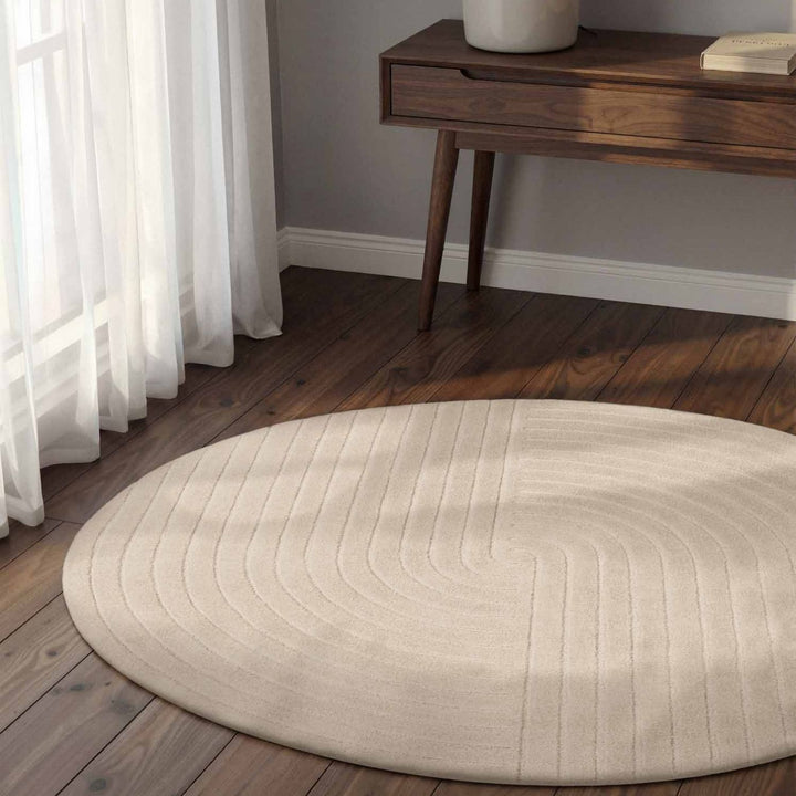 Round beige rug on a wooden floor with a wooden side table and white curtains.