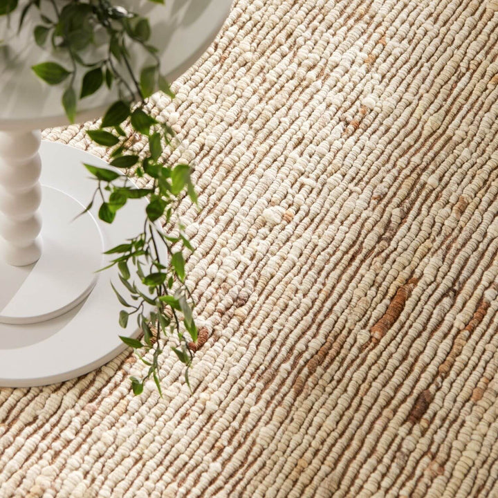 Close-up of a textured beige rug with a white candle holder and green plant in the background.