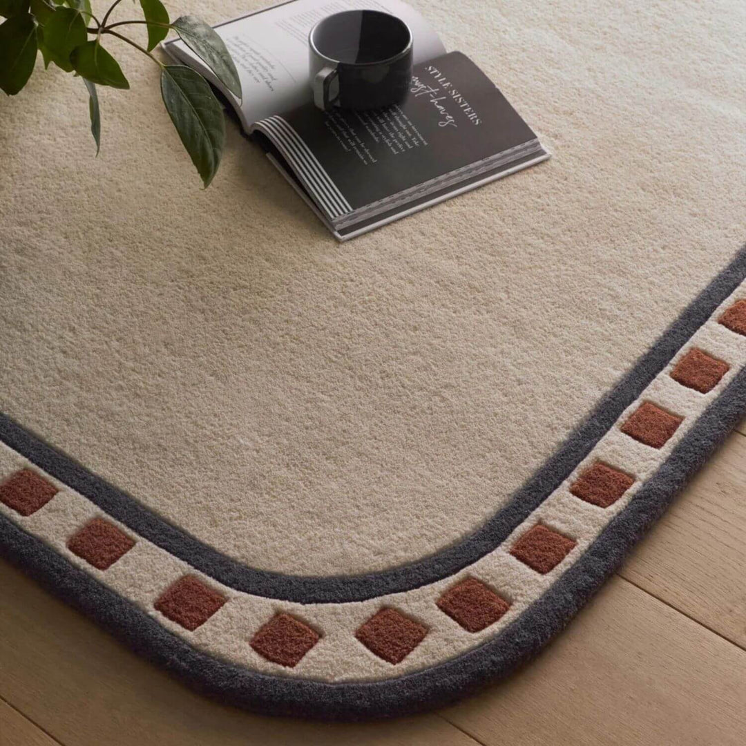 Decorative rug with a geometric border on a wooden floor, featuring a book and cup.