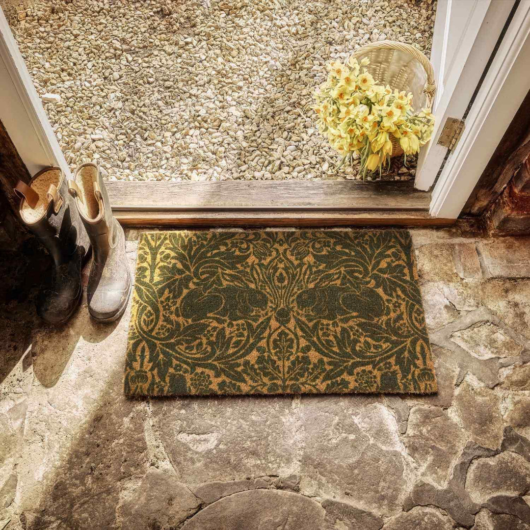 Decorative doormat with floral design on a stone floor, with shoes and flowers in the background.