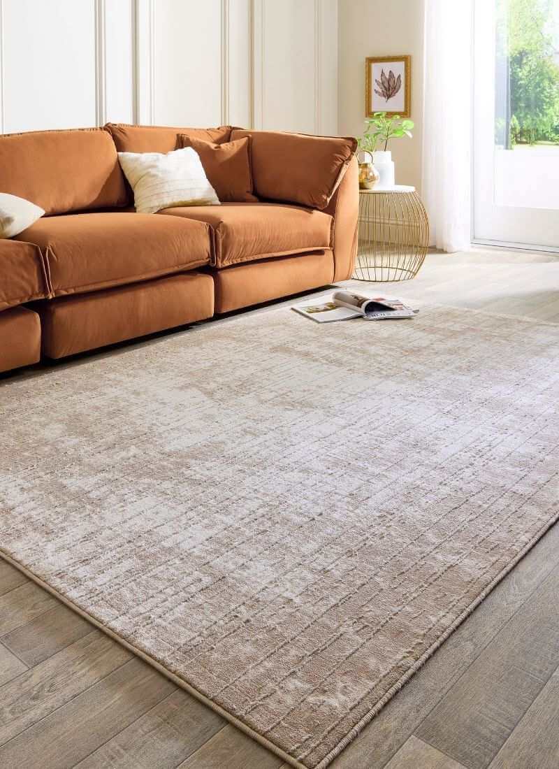 Abstract geometric rug in neutral tones of beige and grey, placed under a cozy orange sofa with soft pillows.