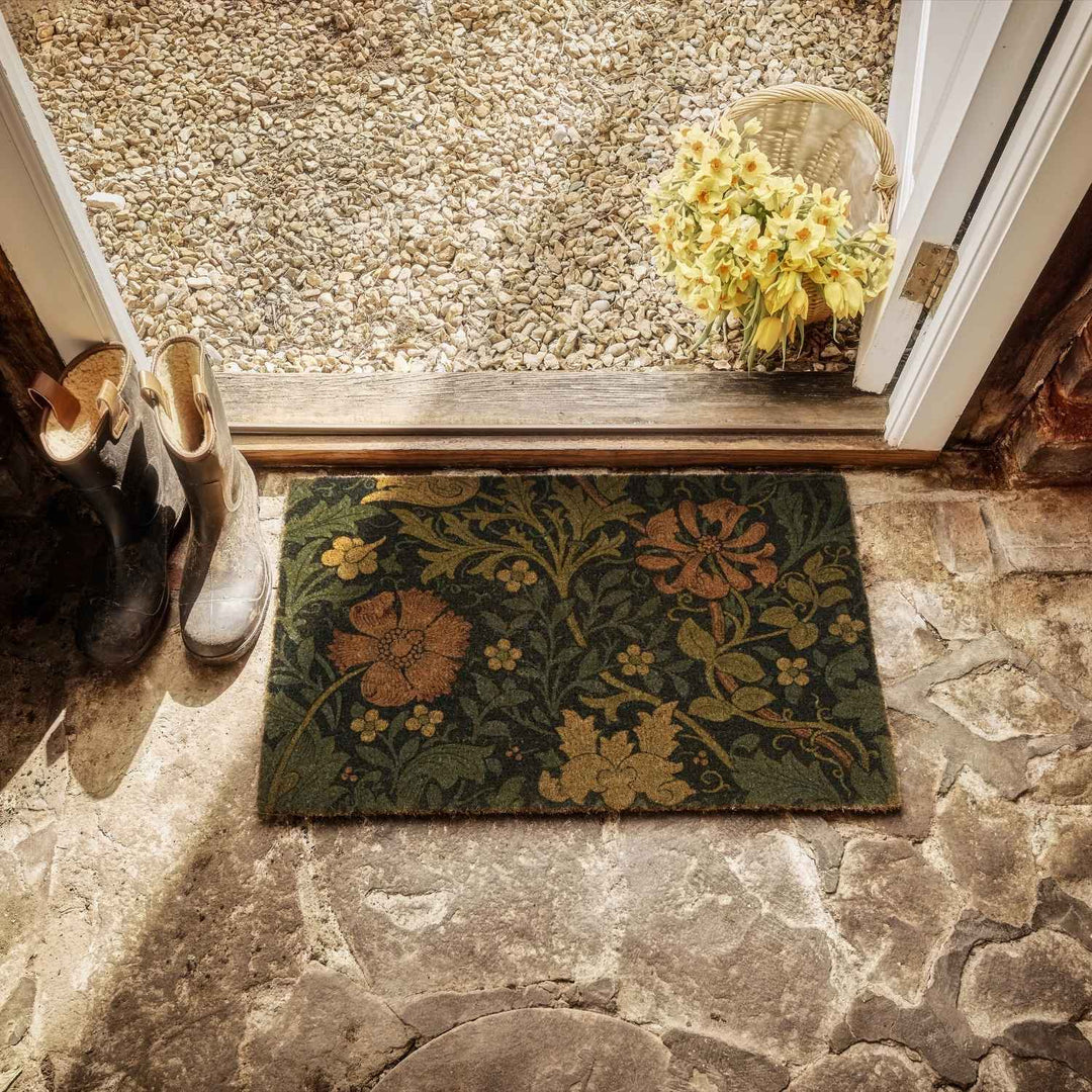Decorative doormat with floral pattern on stone floor, next to shoes and a basket of flowers.