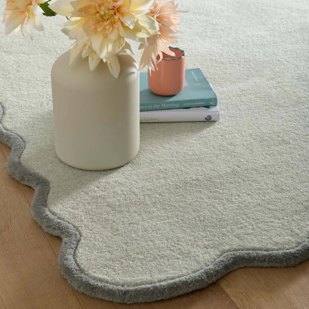 Light green rug with scalloped edge on a wooden floor, featuring a vase with flowers and books.