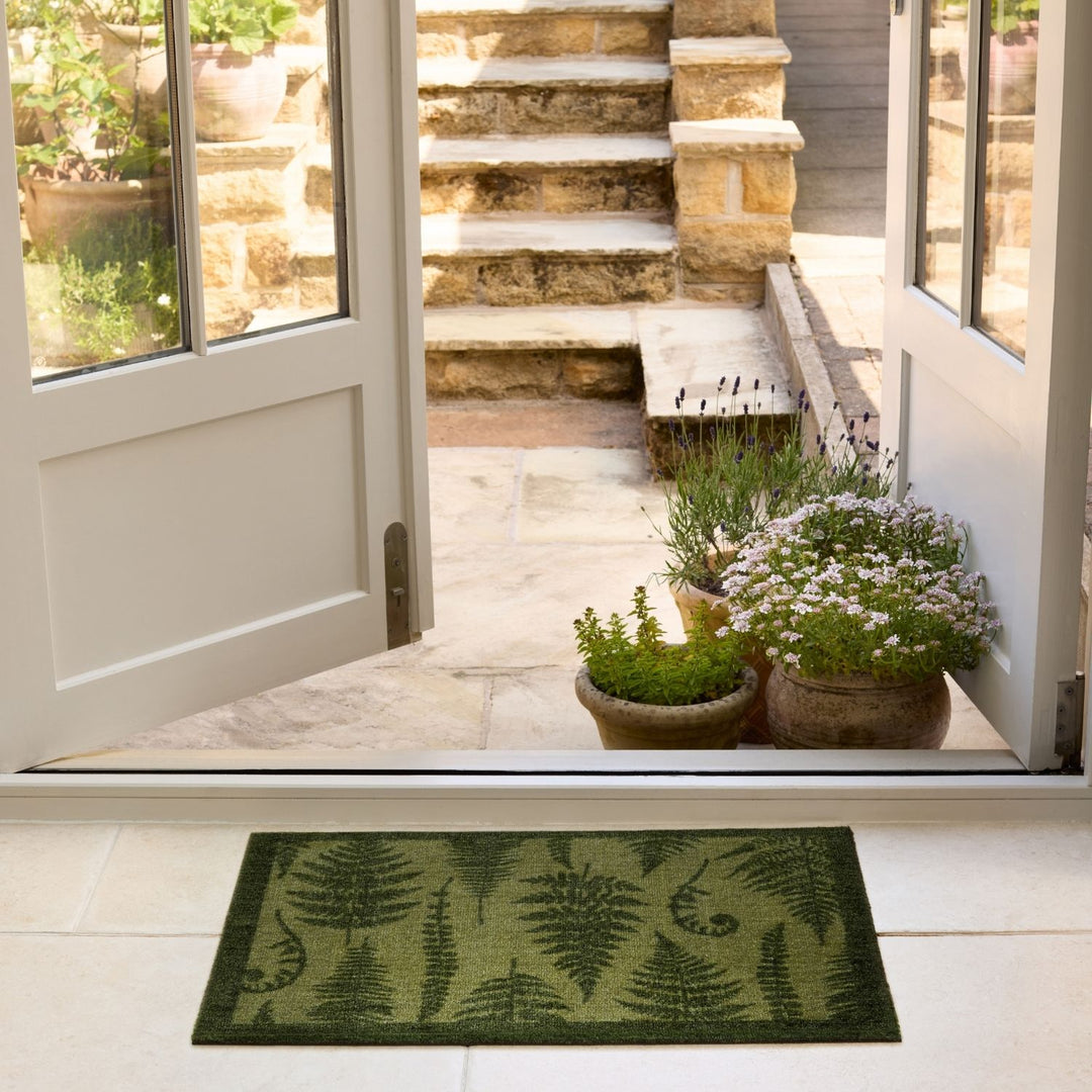 Green doormat with fern pattern in front of a glass door leading to a stone staircase.