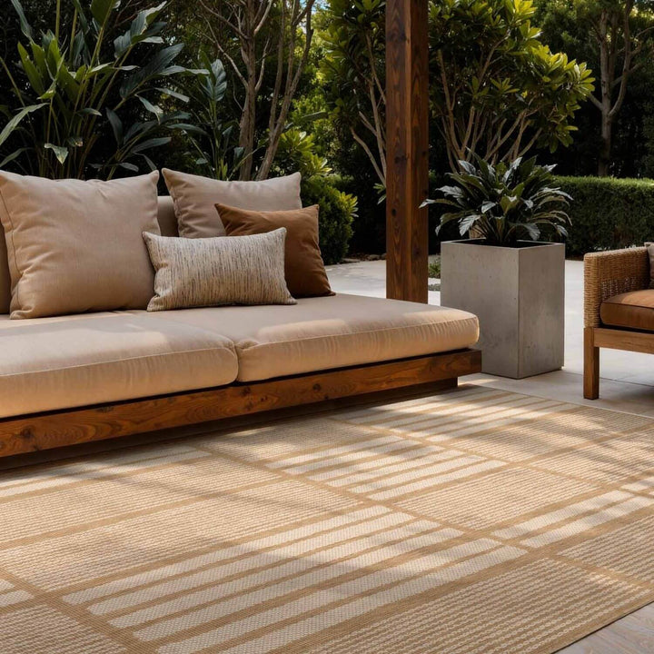 Outdoor seating area with a beige sofa and cushions on a patterned rug, surrounded by greenery.