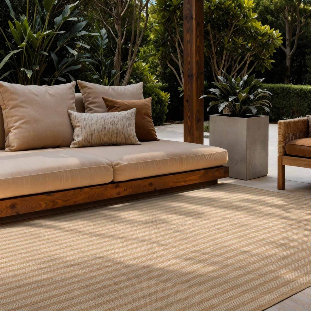 Outdoor seating area with a beige sofa and cushions on a striped rug, surrounded by greenery.