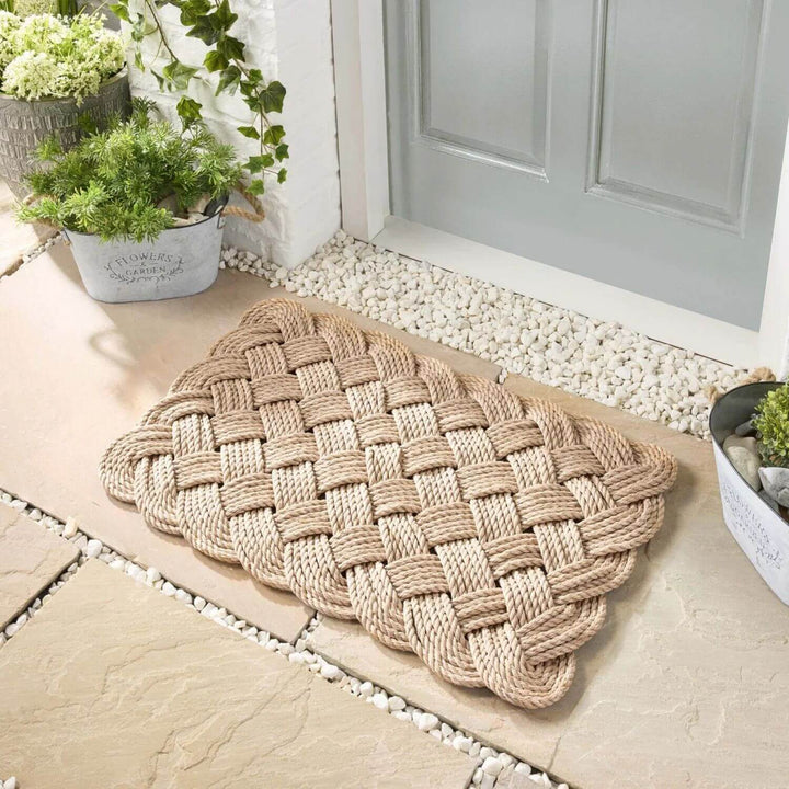 Woven doormat on a stone patio with plants and a door in the background