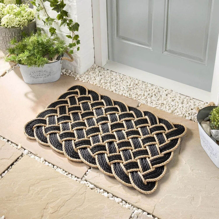 Woven doormat on a stone patio with plants and a door in the background