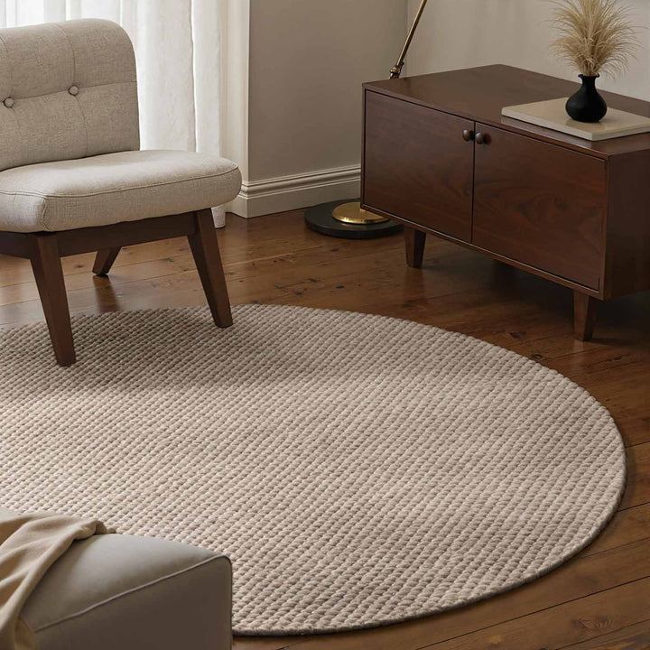 Round beige rug in a living room with a chair and wooden cabinet.