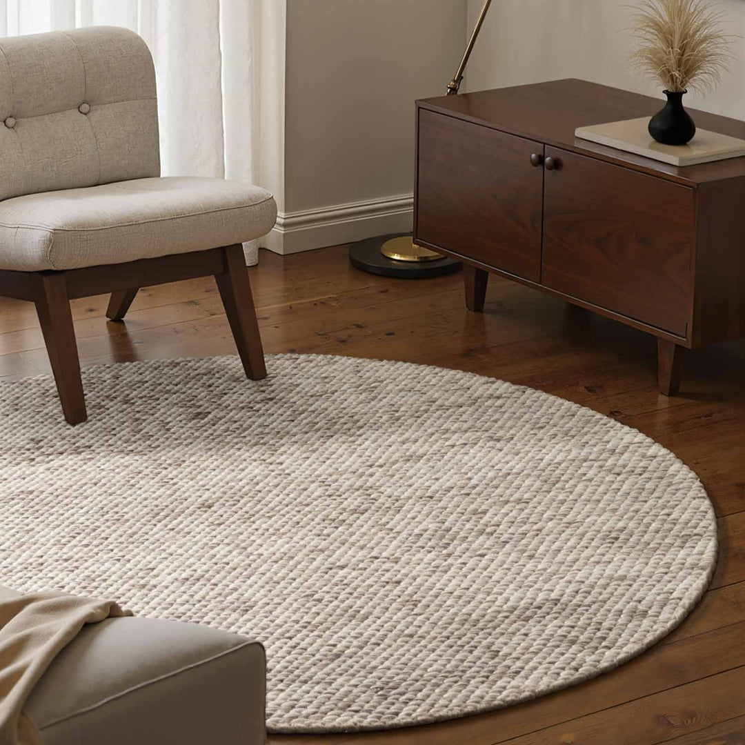 Round beige rug in a living room with a wooden cabinet and chair.