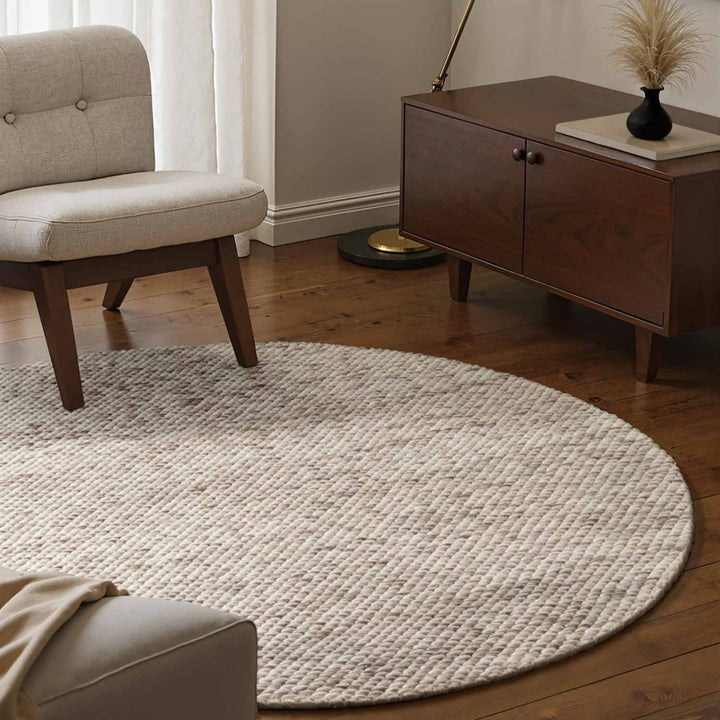 Round beige rug in a living room with a wooden cabinet and chair.