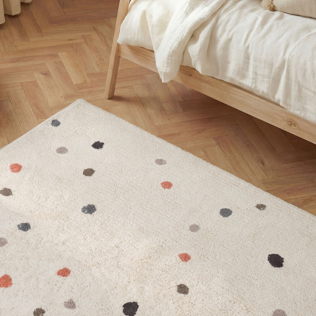 Beige rug with multicolored dots on a wooden floor next to a bed.