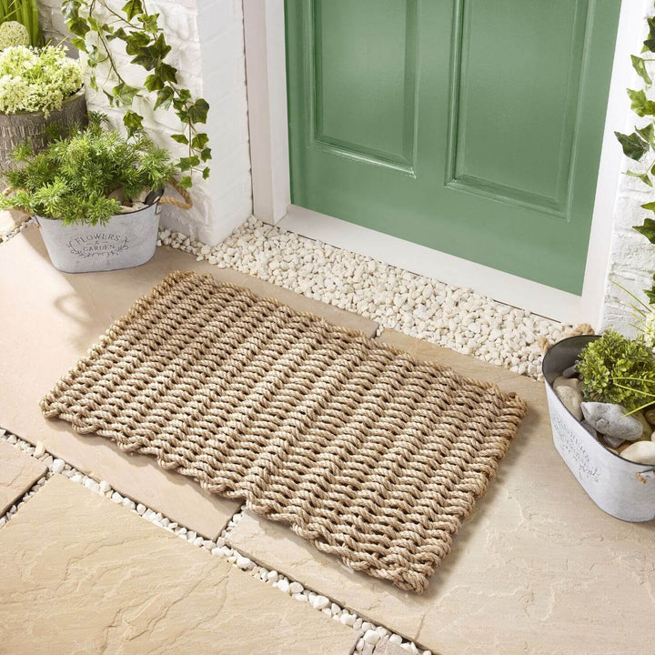 Woven doormat on a stone pathway leading to a green door with plants around.