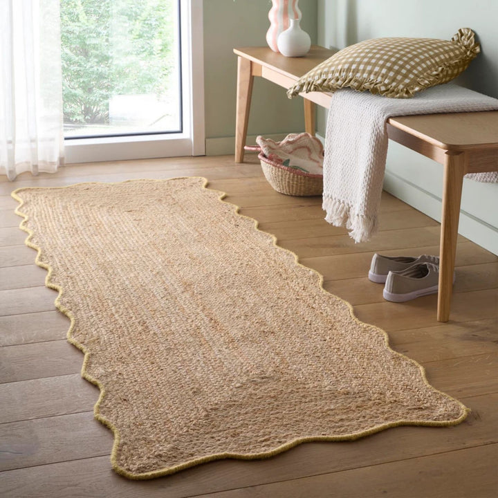 Beige woven rug on a wooden floor with a bench and pillows in the background.