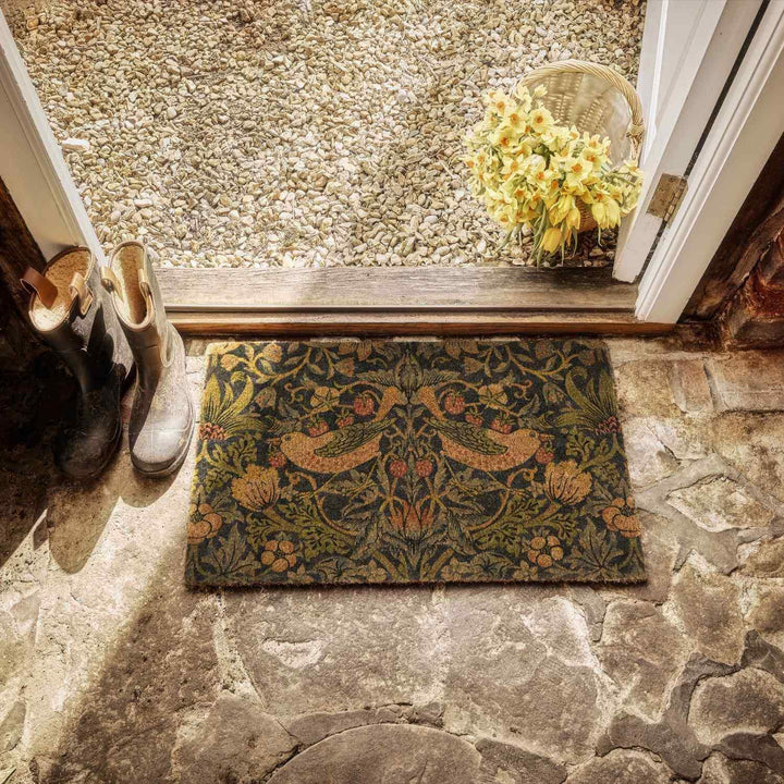 Decorative doormat with floral pattern on stone floor, shoes to the left, basket with flowers above.