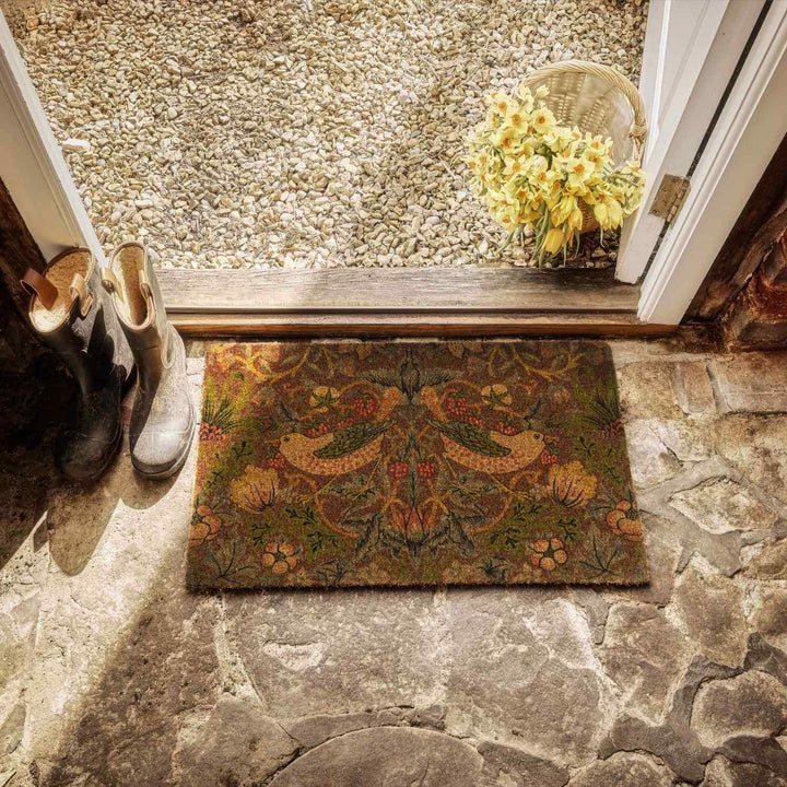 Decorative doormat with a floral pattern on a stone floor, with shoes and a basket of flowers nearby.