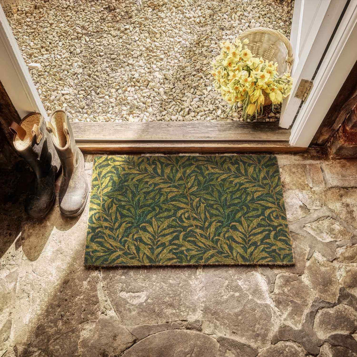 Doormat with leaf pattern on stone floor, shoes, and basket with flowers.