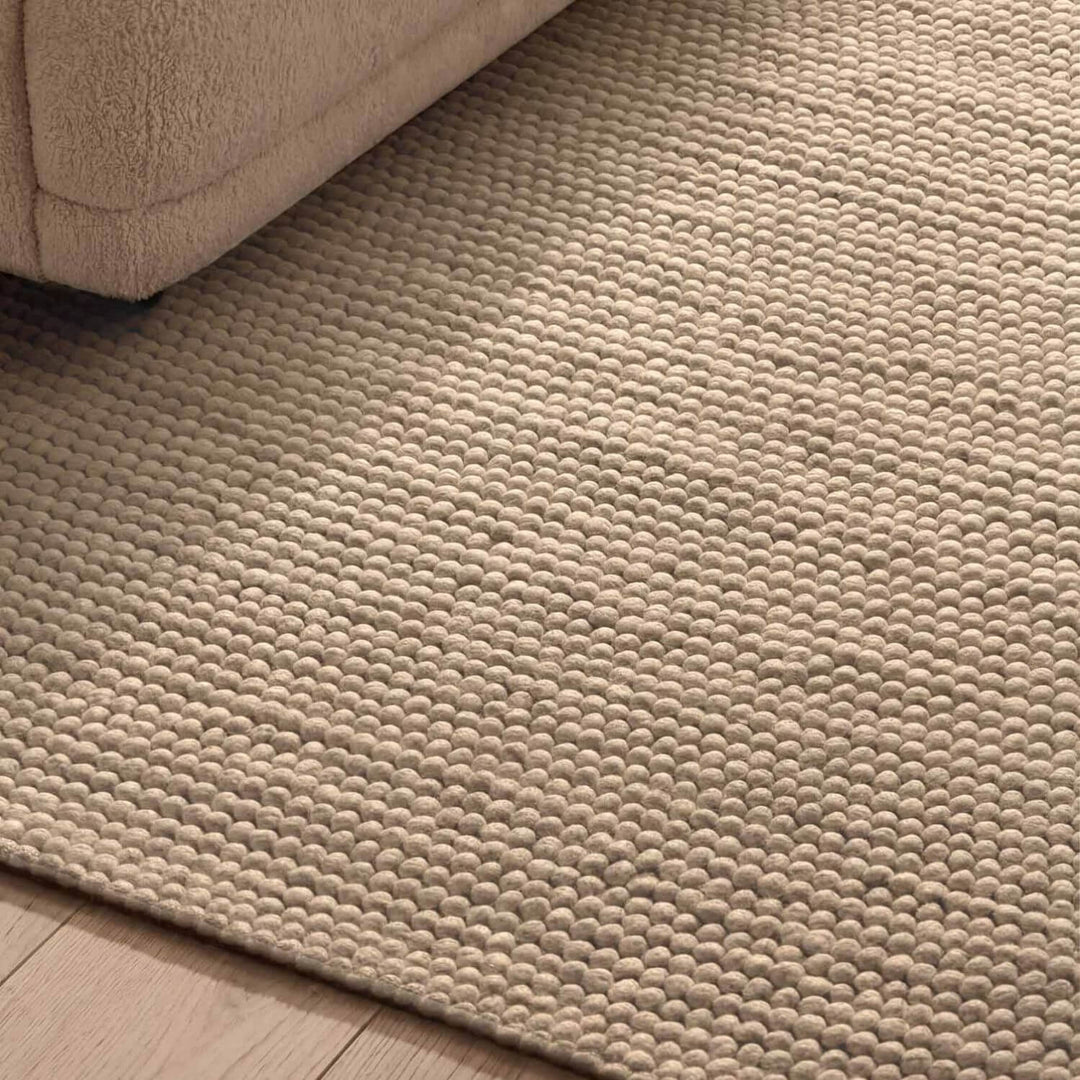 Beige textured rug on a wooden floor with a corner of a beige sofa visible.