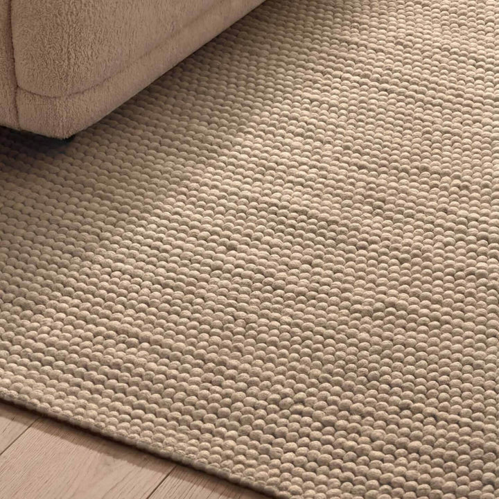 Beige textured rug on a wooden floor with a corner of a beige sofa visible.