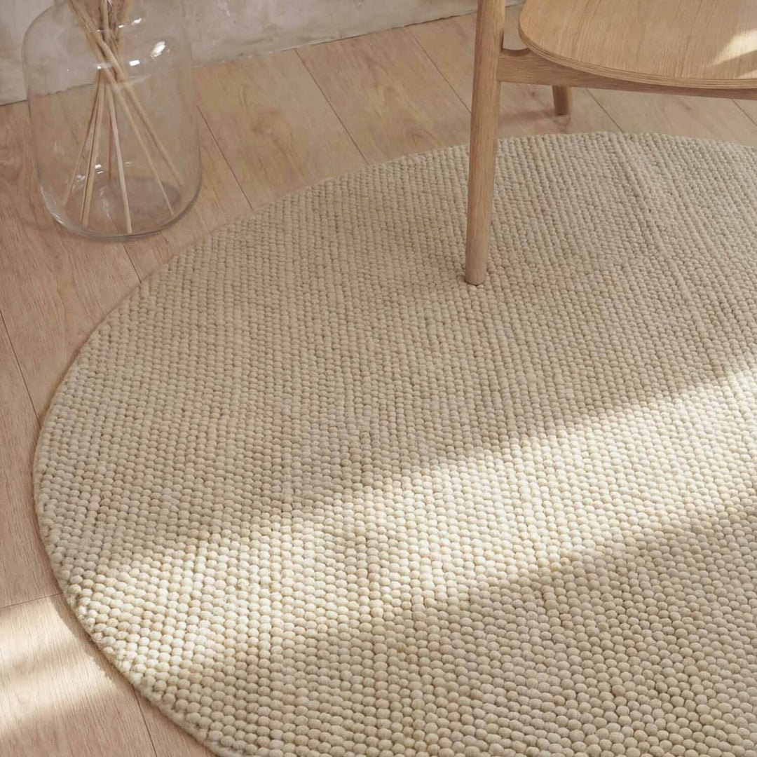 Round beige rug on a wooden floor with a glass jar and wooden chair.