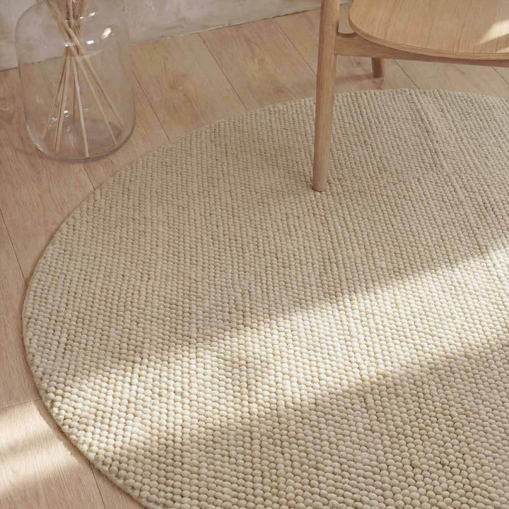 Round beige rug on a wooden floor with a glass jar and wooden chair.