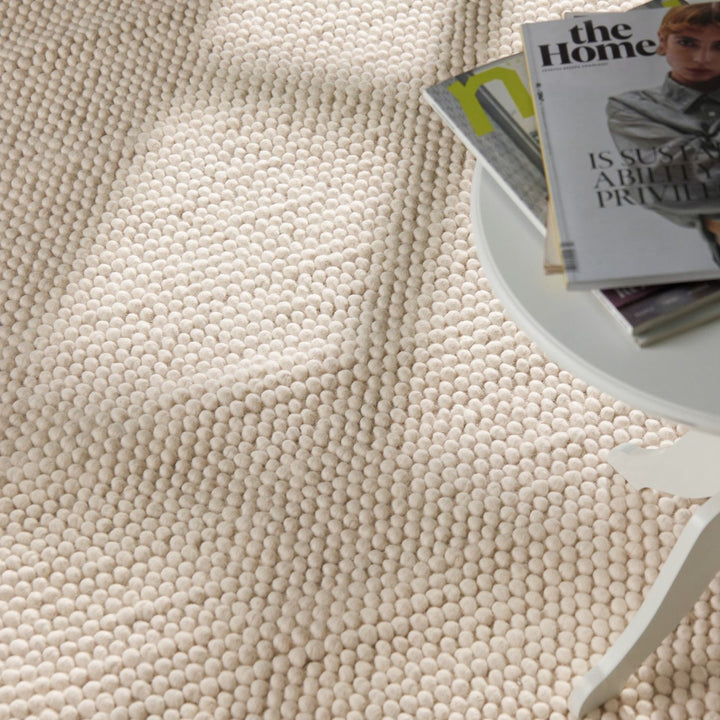 Close-up of a textured beige carpet with a white table and magazines in the background.