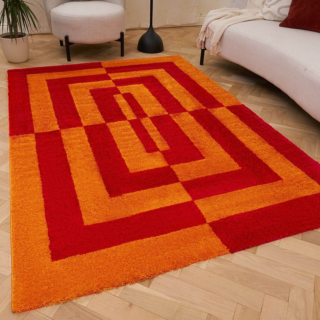 Red and orange geometric rug on a wooden floor with a white sofa and plant in the background.