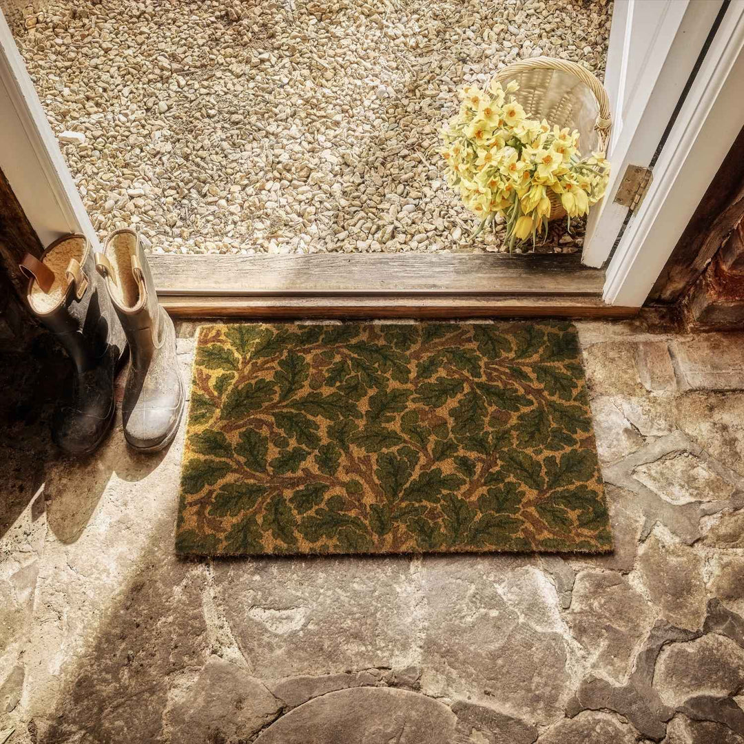 Doormat with leaf pattern on stone floor, shoes, and basket of flowers.