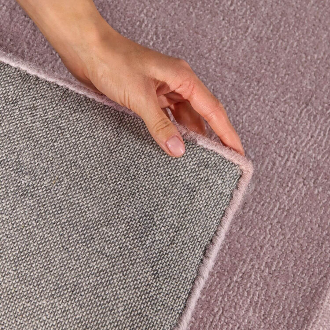 Two hands holding a corner of a gray rug with a pink border.